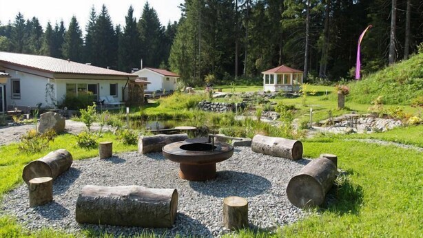 A rustic outdoor area with a fire pit, logs, and a small building. - Haus am Waldpark (Scheidegg)