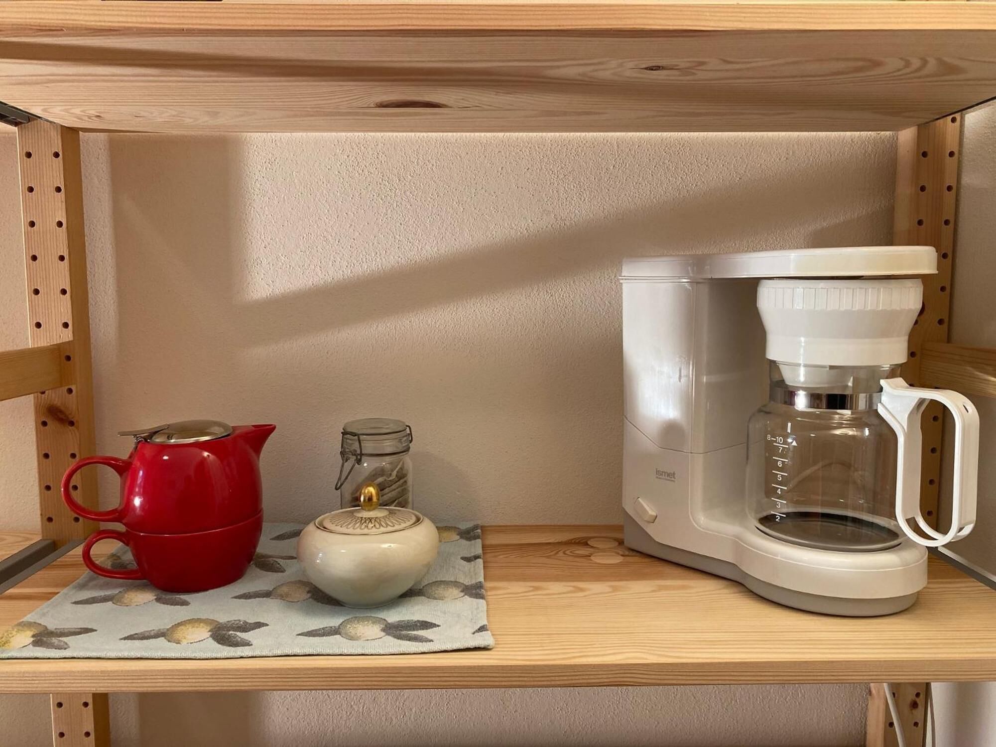 Una estantería de cocina con una cafetera, una tetera roja y un tarro de vidrio.