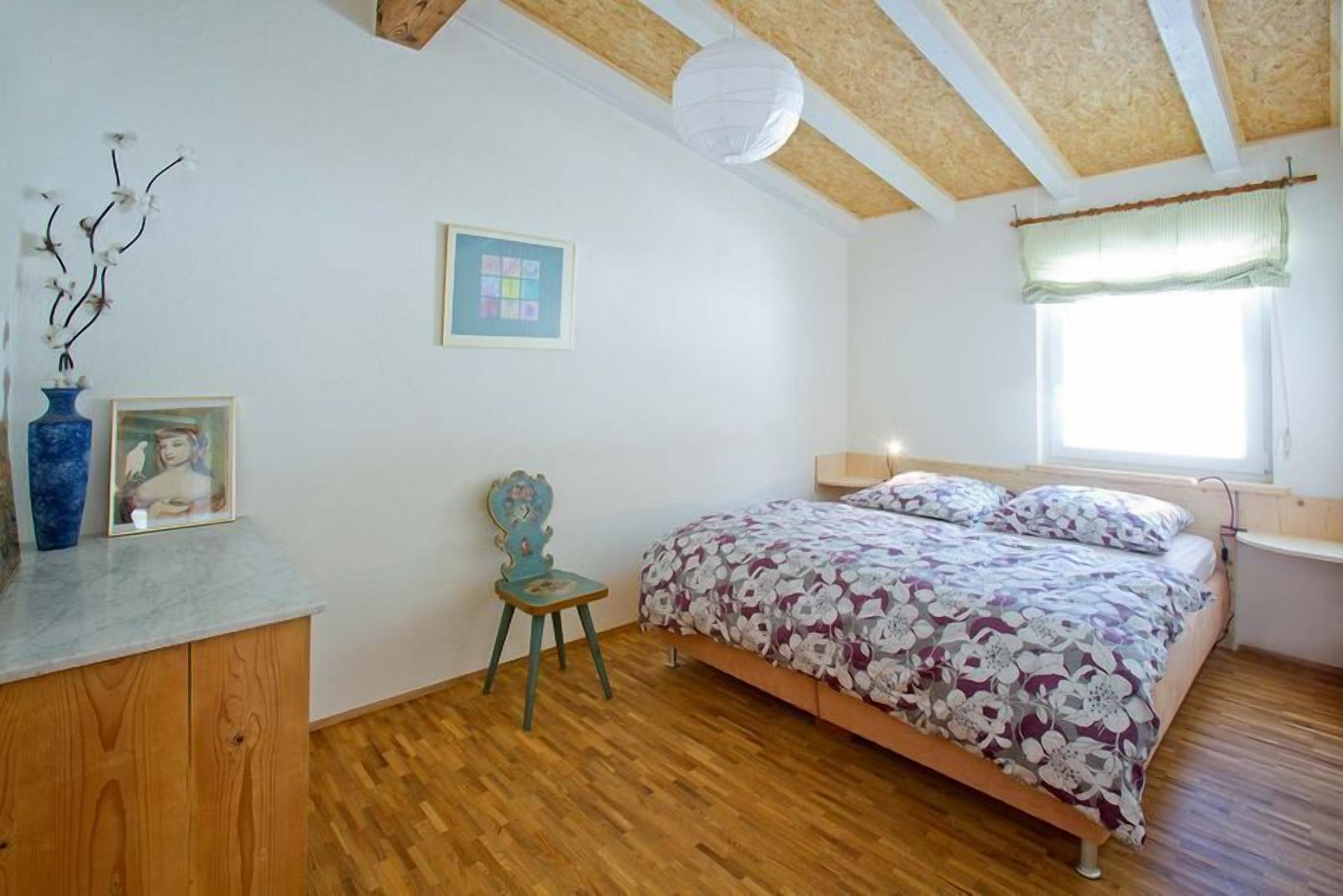 Ein Schlafzimmer mit Holzboden, einem Bett mit Blumenmuster-Bettwäsche, einem Sessel, einem Schreibtisch mit einer Vase und einem gerahmten Bild.