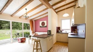 A modern kitchen with wooden beams, a dining area with a red sofa, and a large window overlooking a garden. - Haus am Waldpark (Scheidegg)