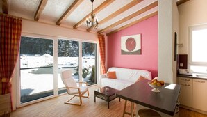 A cozy room with a wooden ceiling, a white sofa, a dining table with a fruit bowl, and a view of a snowy landscape through large windows. - Haus am Waldpark (Scheidegg)