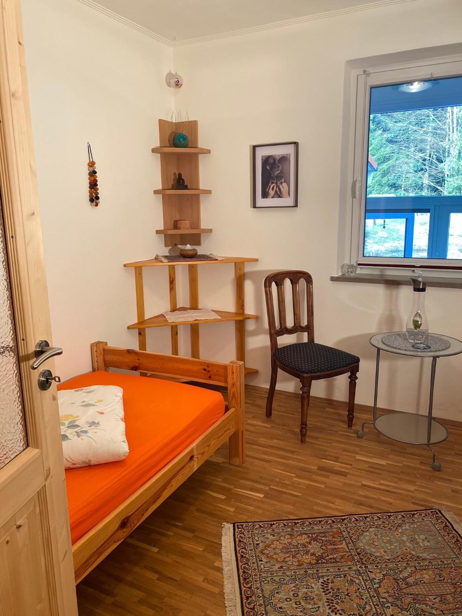 Pieni, viihtyisä huone, jossa on puinen sänky, tuoli, pieni pöytä, koriste-esineillä täytetty hylly ja ikkuna, josta avautuu näkymä puihin.