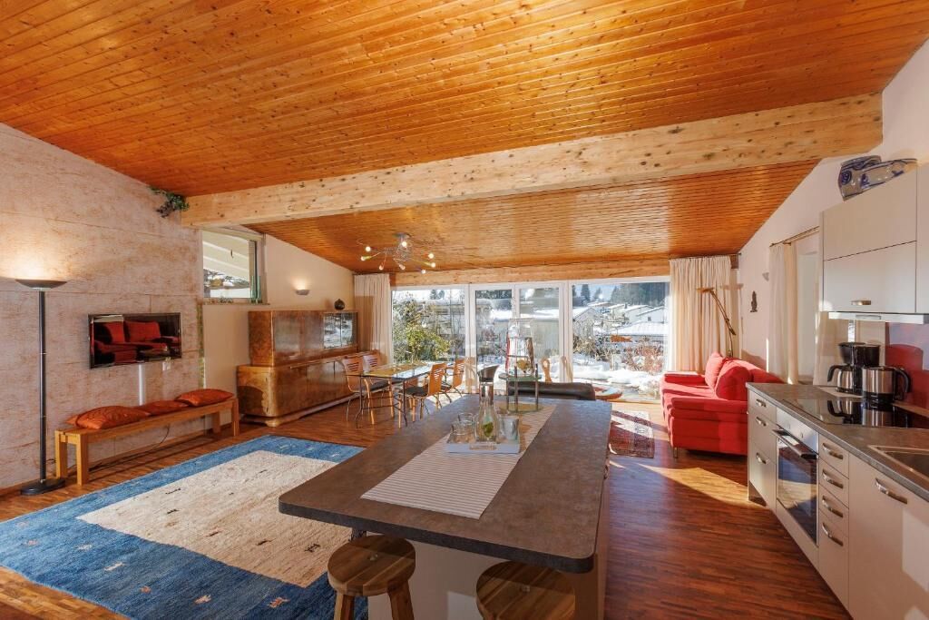 Amplia sala de estar con techo de madera, cocina equipada con electrodomésticos modernos y un ventanal con vistas al exterior.
