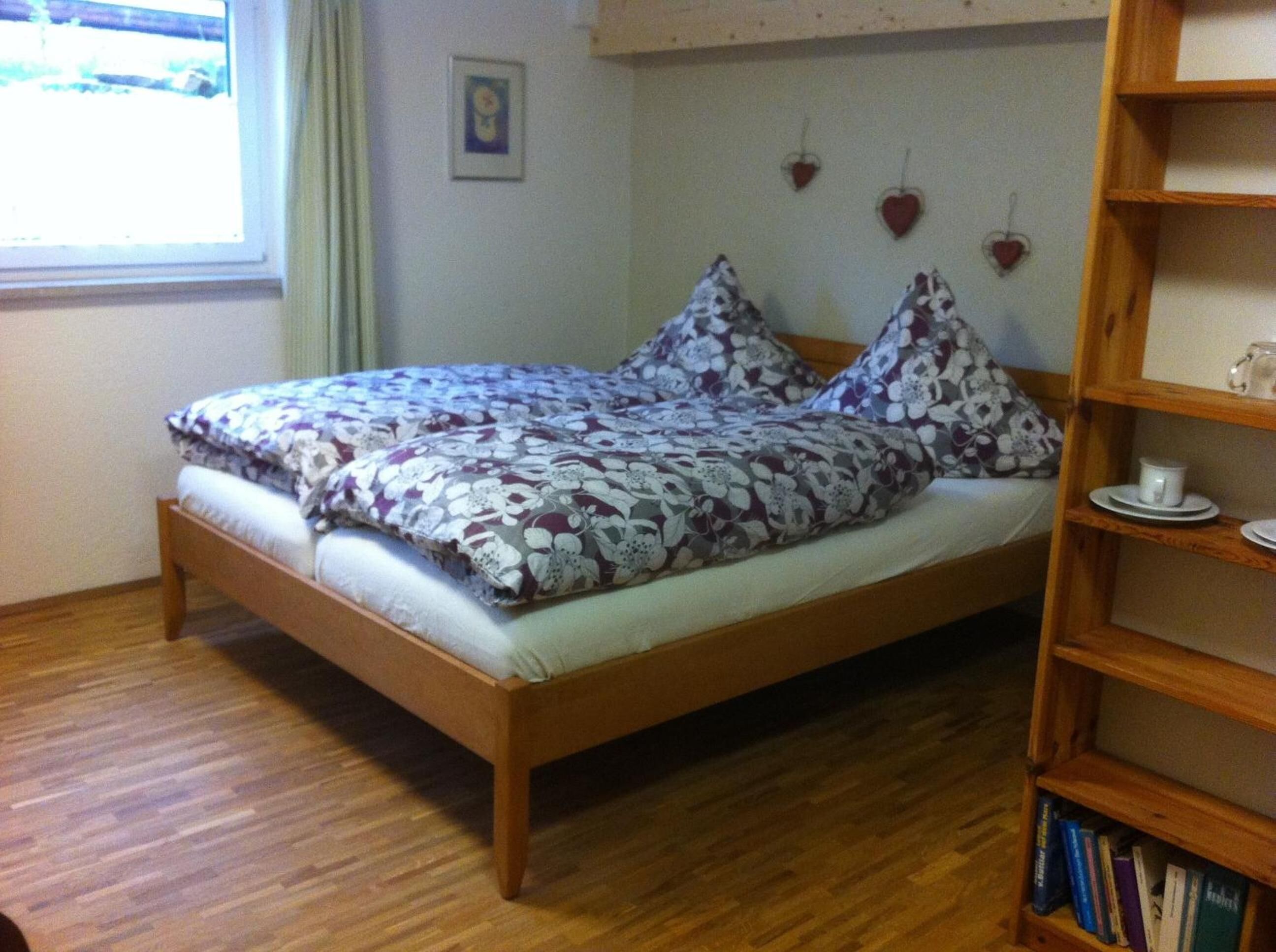 A bed with floral bedding, a wooden headboard, a bedside shelf with books, and a window with curtains.