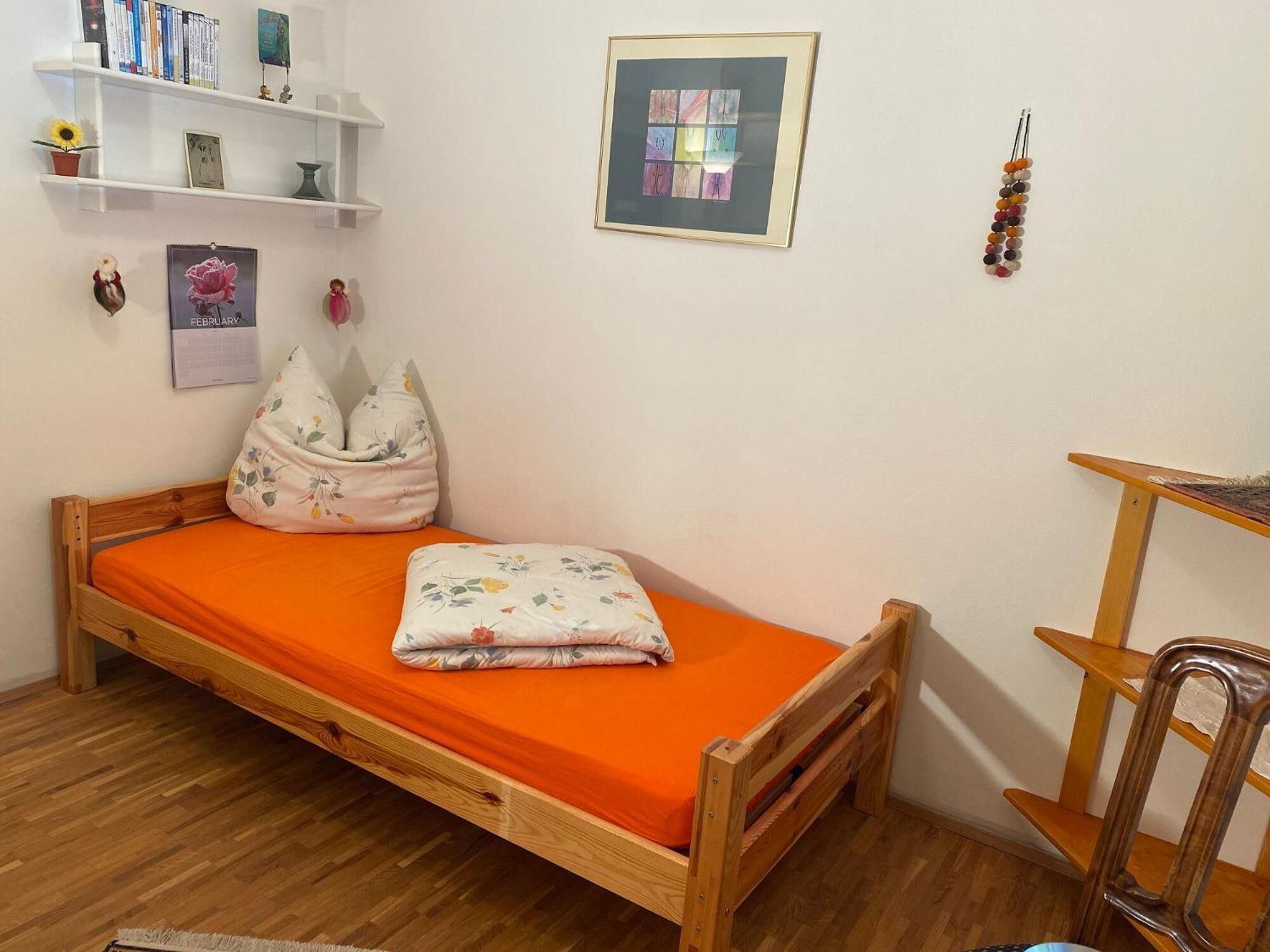 Une petite chambre à coucher, bien rangée, avec un lit en bois, une literie orange, une petite étagère avec des livres et des objets décoratifs, un tableau abstrait encadré et un collier de perles accroché au mur.