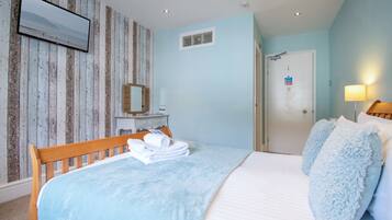 A bedroom with a wooden headboard, a bed with a blue blanket, two fluffy pillows, a wooden dresser, a mirror, and a television mounted on the wall.