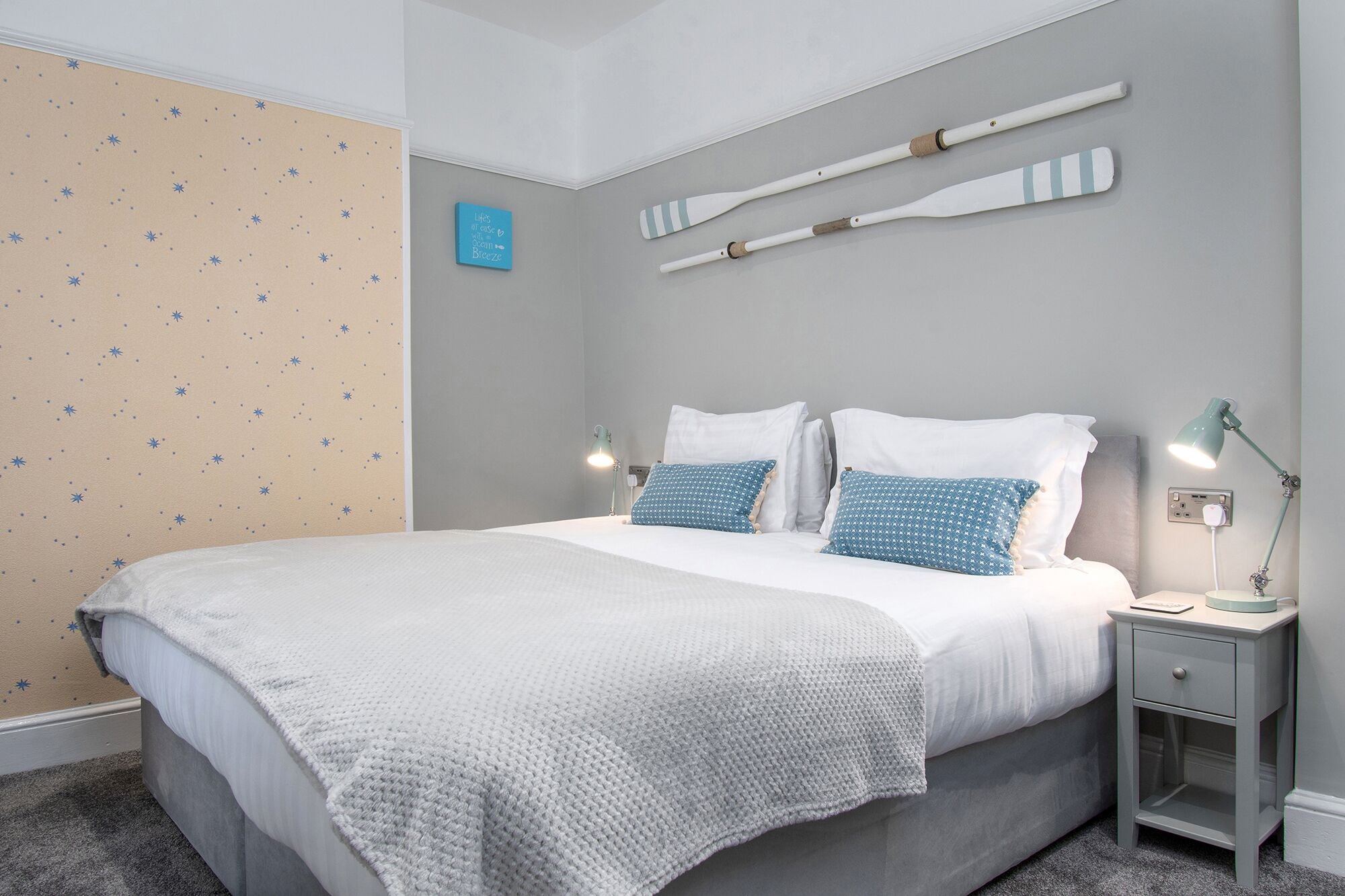 Una cama bien hecha con ropa de cama blanca y azul, una mesita de noche pequeña con una lámpara, y una pared con papel tapiz estampado con estrellas y dos palas de madera.