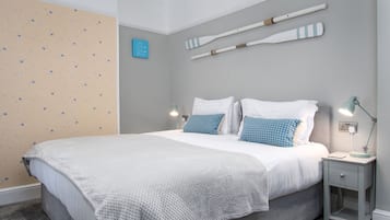Una cama bien hecha con ropa de cama blanca y azul, una mesita de noche pequeña con una lámpara, y una pared con papel tapiz estampado con estrellas y dos palas de madera.
