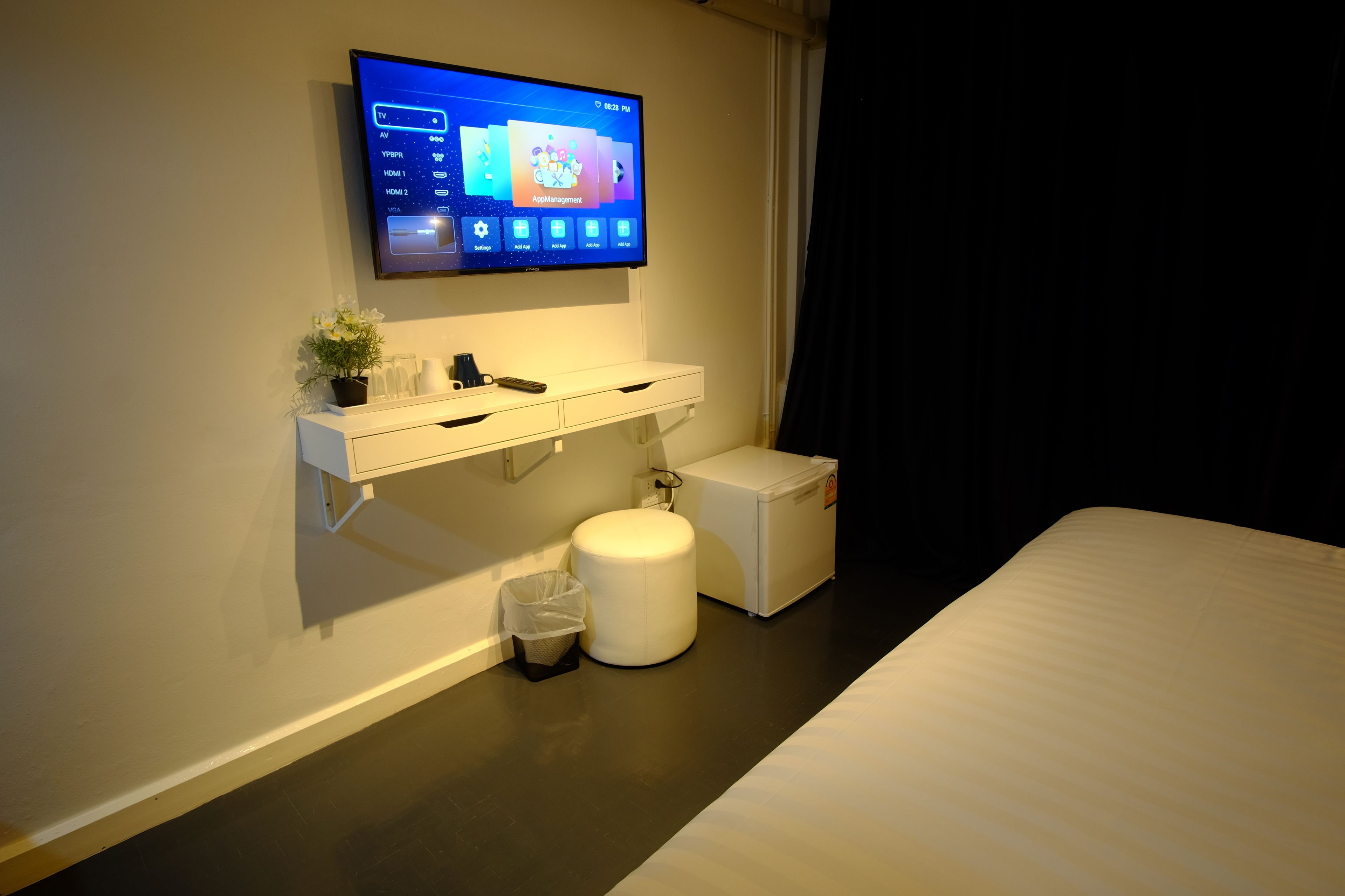 A hotel room with a mounted TV displaying a home screen, a small potted plant, a white bedside table with a remote control, a trash bin, and a white bedside unit.