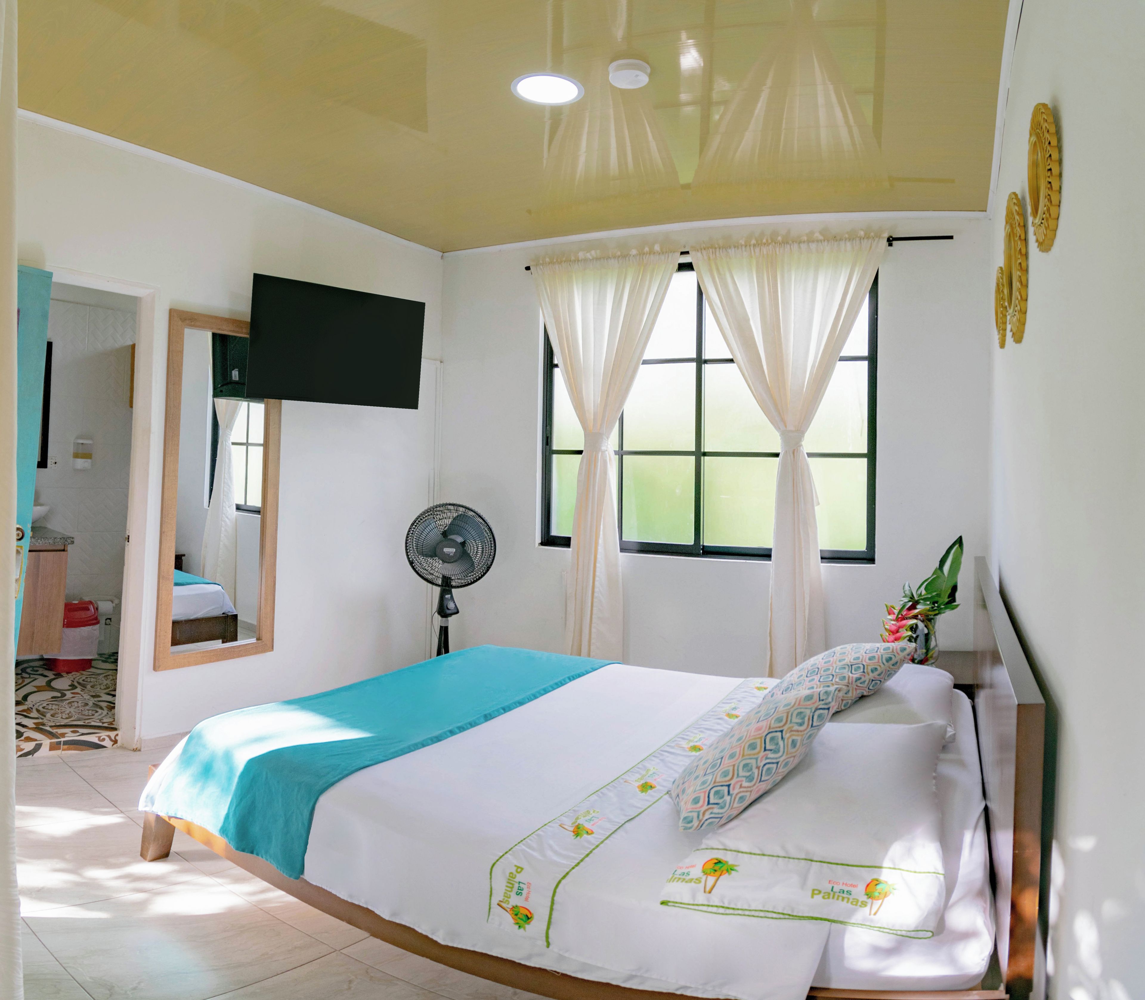 Une chambre d’hôtel avec un lit, un téléviseur fixé au mur, une fenêtre avec des rideaux, un ventilateur sur pied et une salle de bain visible à travers une porte ouverte.