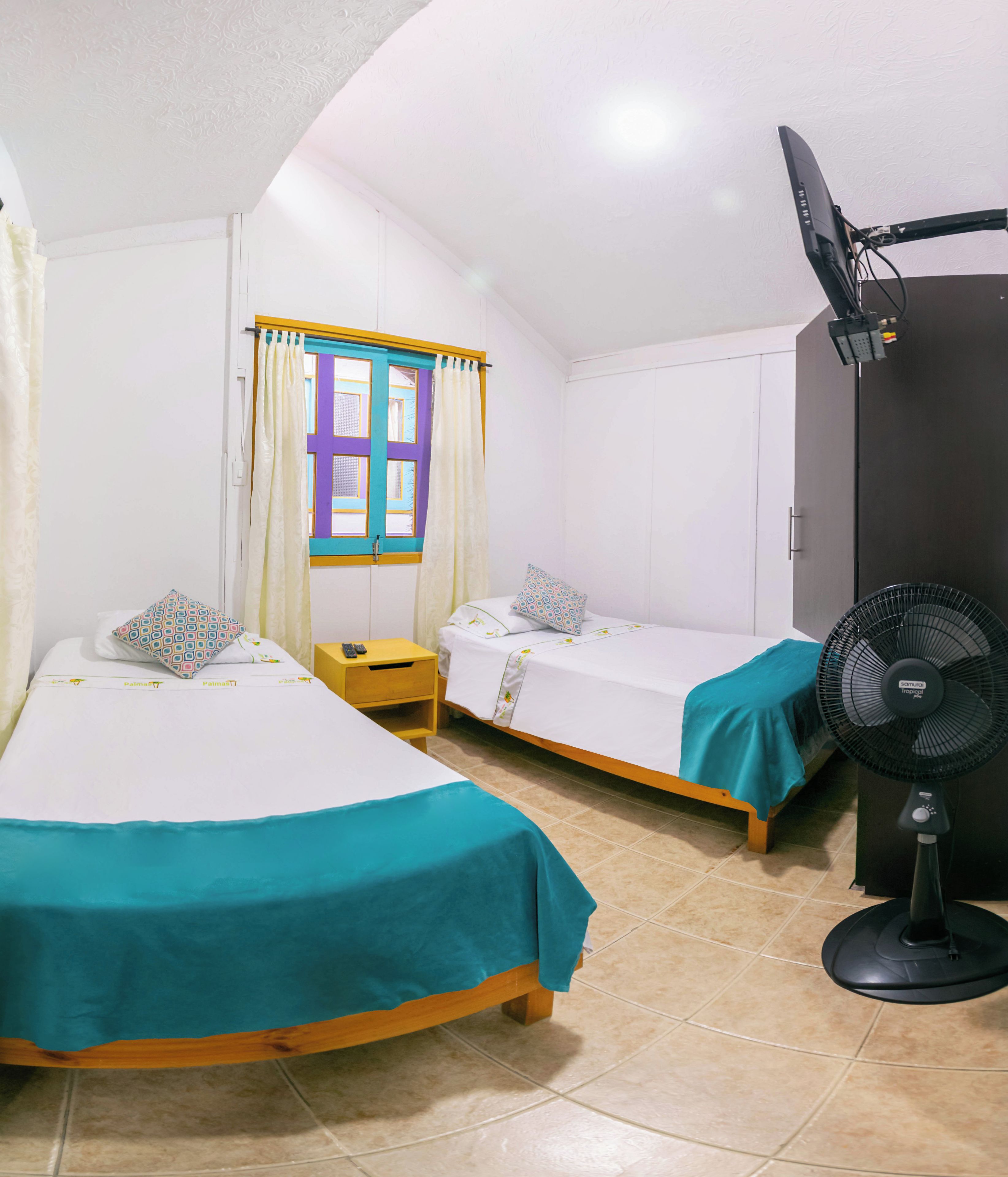 A room with two single beds, a window with curtains, a wardrobe, and a standing fan.