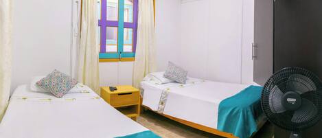 Una habitación con dos camas individuales, una ventana con cortinas, un armario y un ventilador de pie.
