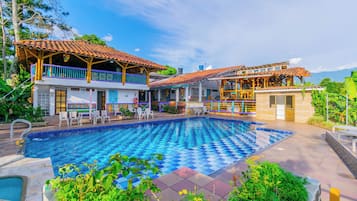 A large outdoor swimming pool with a checkered pattern, surrounded by a patio area with outdoor seating and a view of the mountains.