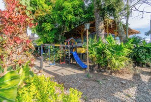 A playground with a slide, surrounded by lush greenery and a wooden structure.