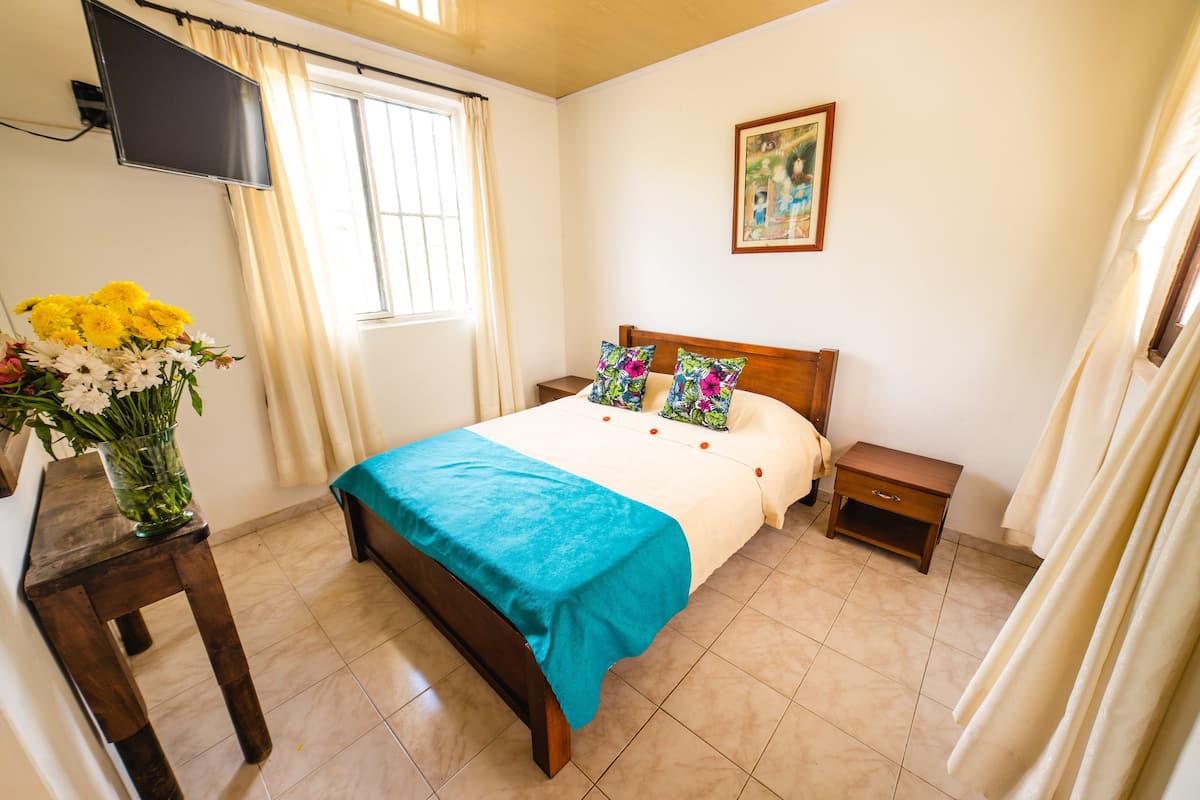 Un dormitorio ordenado con una cama, mesitas de noche, un televisor montado en la pared y una ventana con cortinas.