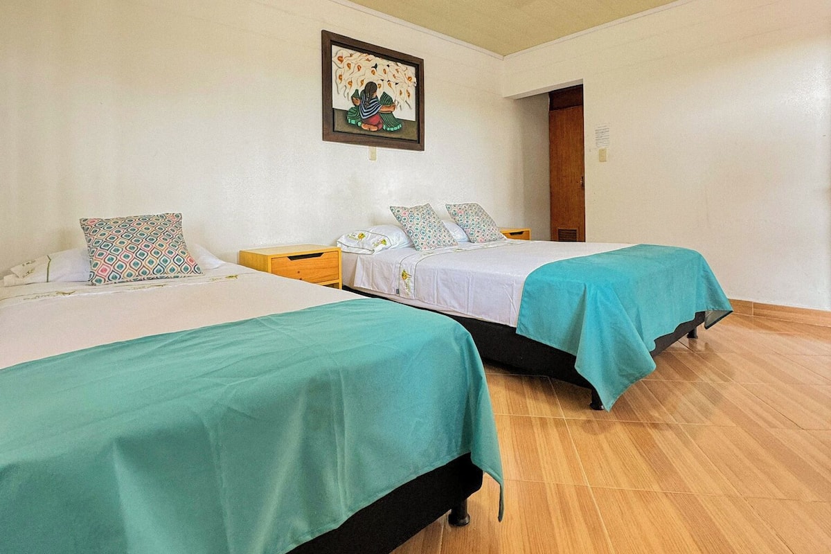 Una habitación con dos camas, cada una con cubrecama color turquesa y ropa de cama blanca. Hay un cuadro enmarcado en la pared sobre las camas.