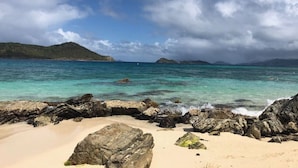 Een strand met helder turquoise water, een rotsachtige kustlijn en verre bergen onder een lichtbewolkte lucht.