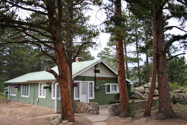 A small, green lodge with a stone entrance, surrounded by tall trees.