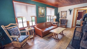 Una sala de estar acogedora con piso de madera, un sofá de cuero marrón, dos sillones de madera con cojines, una mesita de madera y una chimenea.