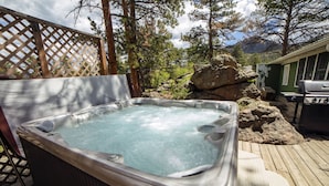 Un jacuzzi en una terraza de madera con vista a árboles y una parrilla.