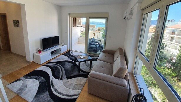 En moderne stue med et fladskærms-tv, en beige sofa og et stort vindue med udsigt til havet og bygninger.