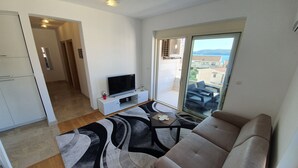 En moderne stue med sofa, et fladskærms-tv på et hvidt skab, et mønstret tæppe og en skydedør, der fører til et udendørs opholdsområde med udsigt til bygninger og bakker.