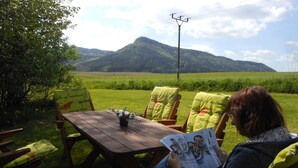 A person reading a magazine at a wooden table outdoors with chairs, a grassy field, and mountains in the background. - Nostalgic renovated farmhouse in beautiful surroundings (Božanov)