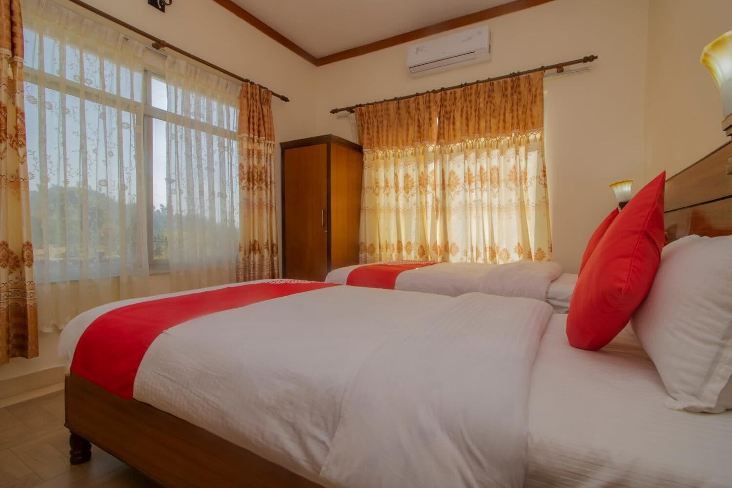 A hotel room with two beds, each with white bedding and red pillows, a wooden wardrobe, and a window with sheer curtains.