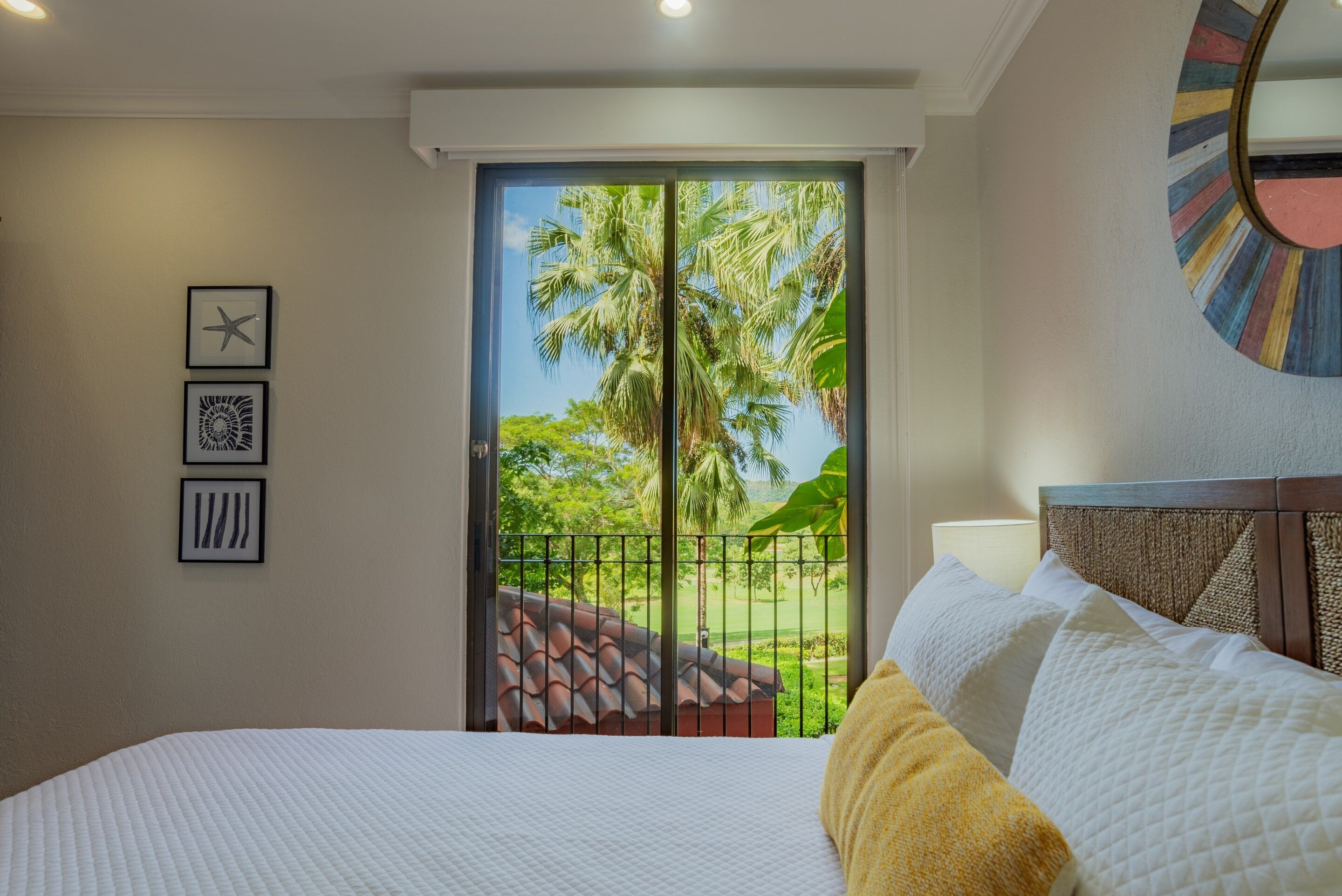 A hotel room with a bed, a view of palm trees, and a balcony with a railing.