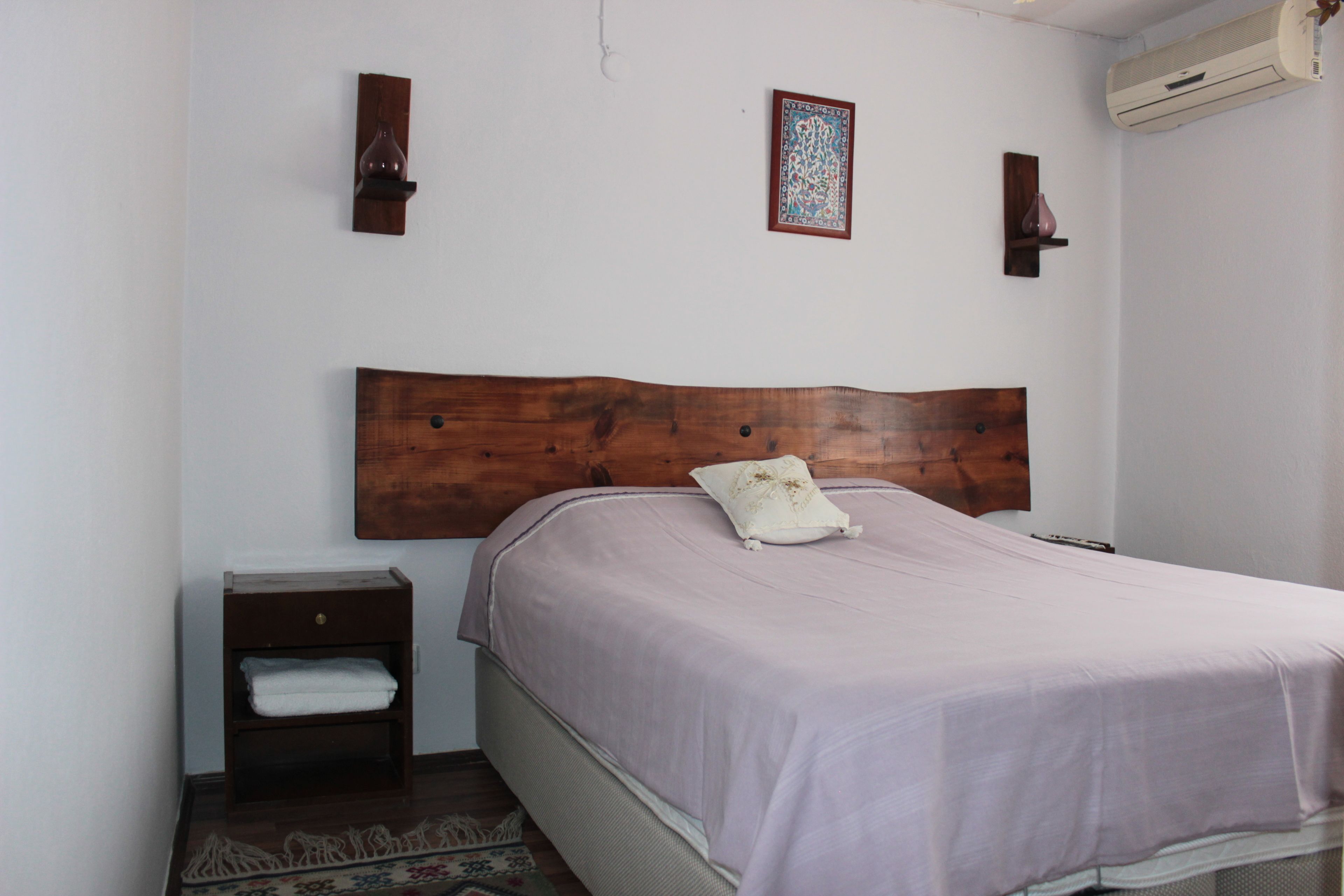 Un dormitorio con cabecera de madera, una cama individual con almohada blanca, una mesita de noche con cajón y una pequeña estantería.
