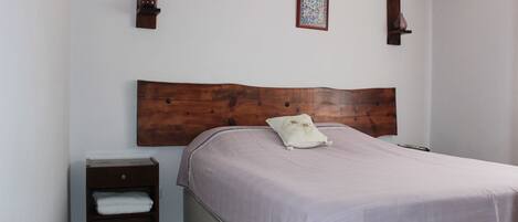 Un dormitorio con cabecera de madera, una cama individual con almohada blanca, una mesita de noche con cajón y una pequeña estantería.