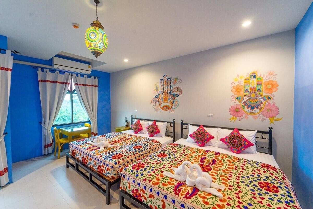 A hotel room with two beds, colorful patterned bedspreads, a small table with a chair, a window with curtains, and decorative wall art.