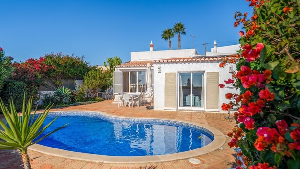 A swimming pool with a tiled deck, a patio area with a table and chairs, and a building with a tiled roof and windows. - Casa Giovanna Praia da Luz 3 bedrooms, aircon, solar heated pool & sea view. (Luz)