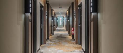 A long, narrow hallway with doors on both sides, numbered signs above each door, and a fire extinguisher on the right wall.