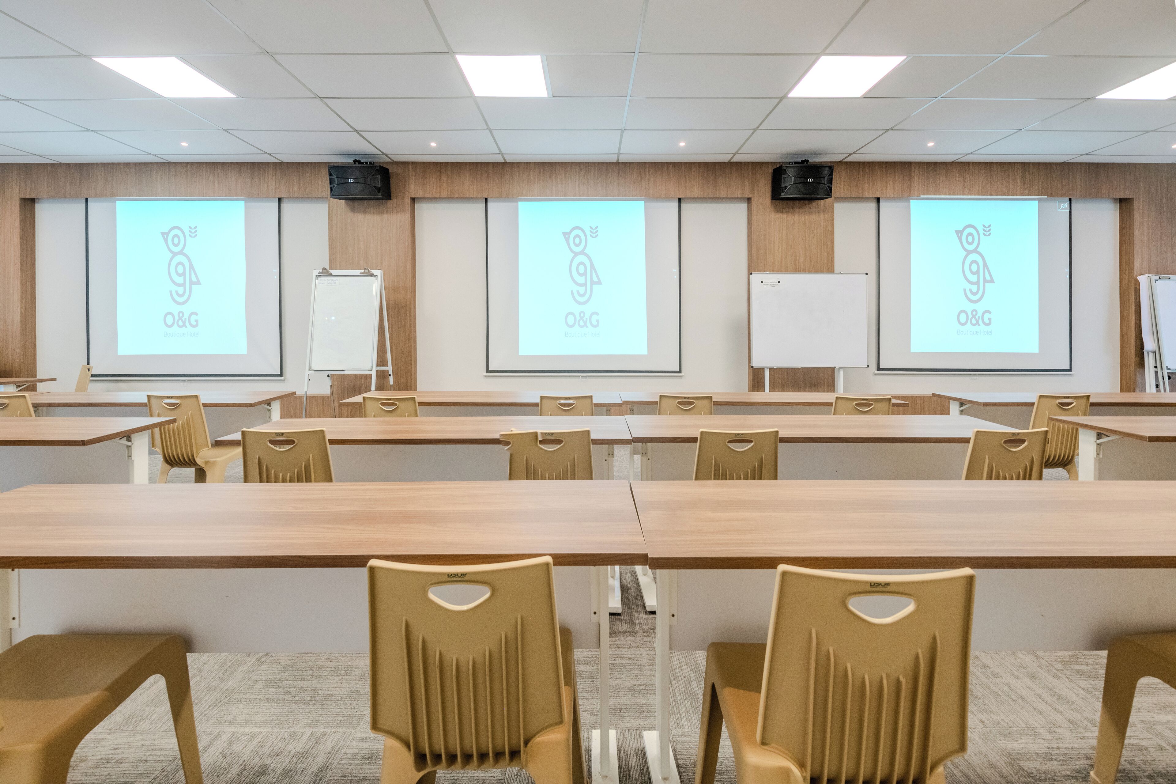 A conference room with long tables and chairs, a whiteboard, and a projector screen displaying the O&G logo.