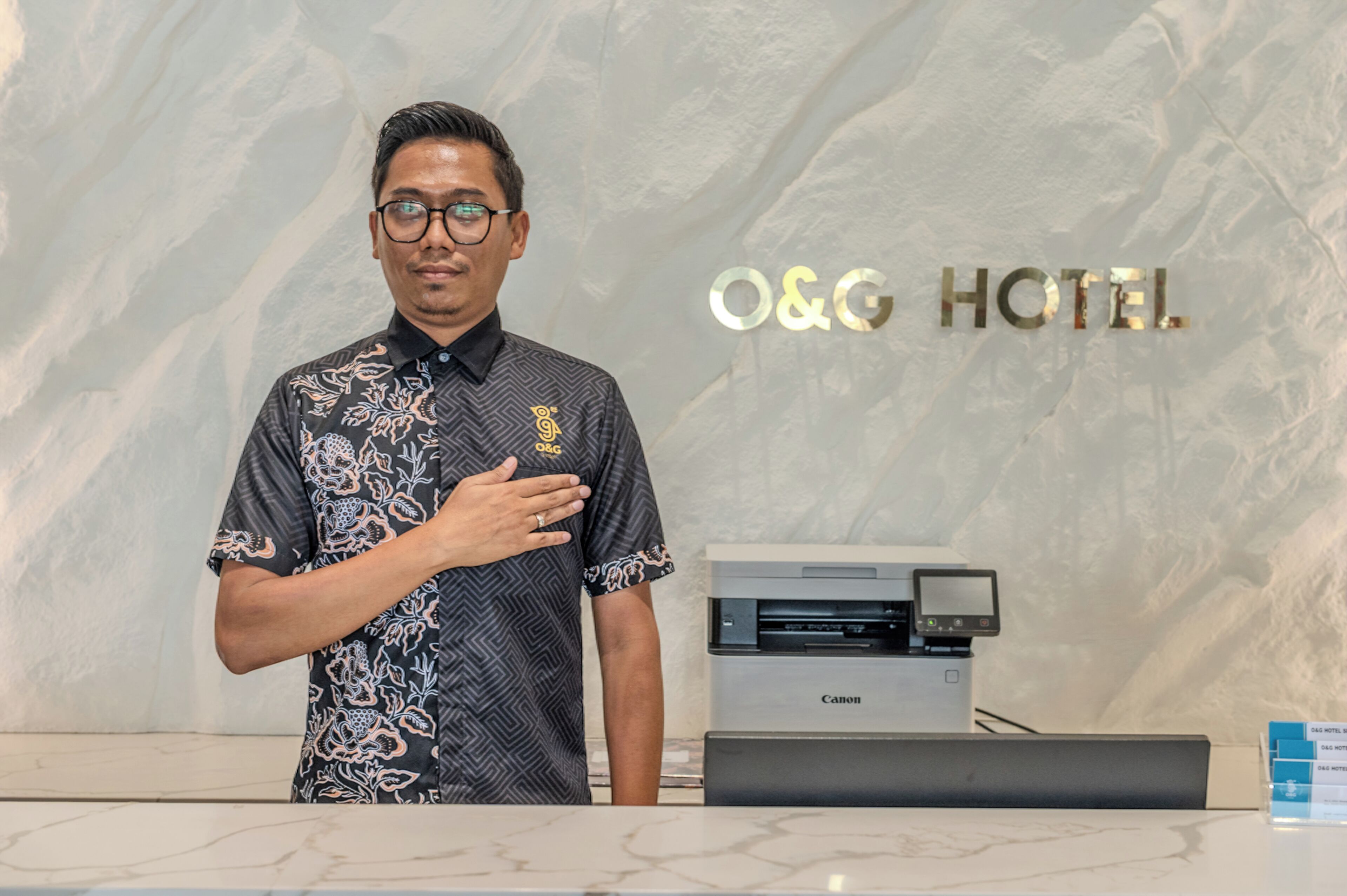 A man standing at a reception desk with a Canon printer and a sign that reads "O&G HOTEL".