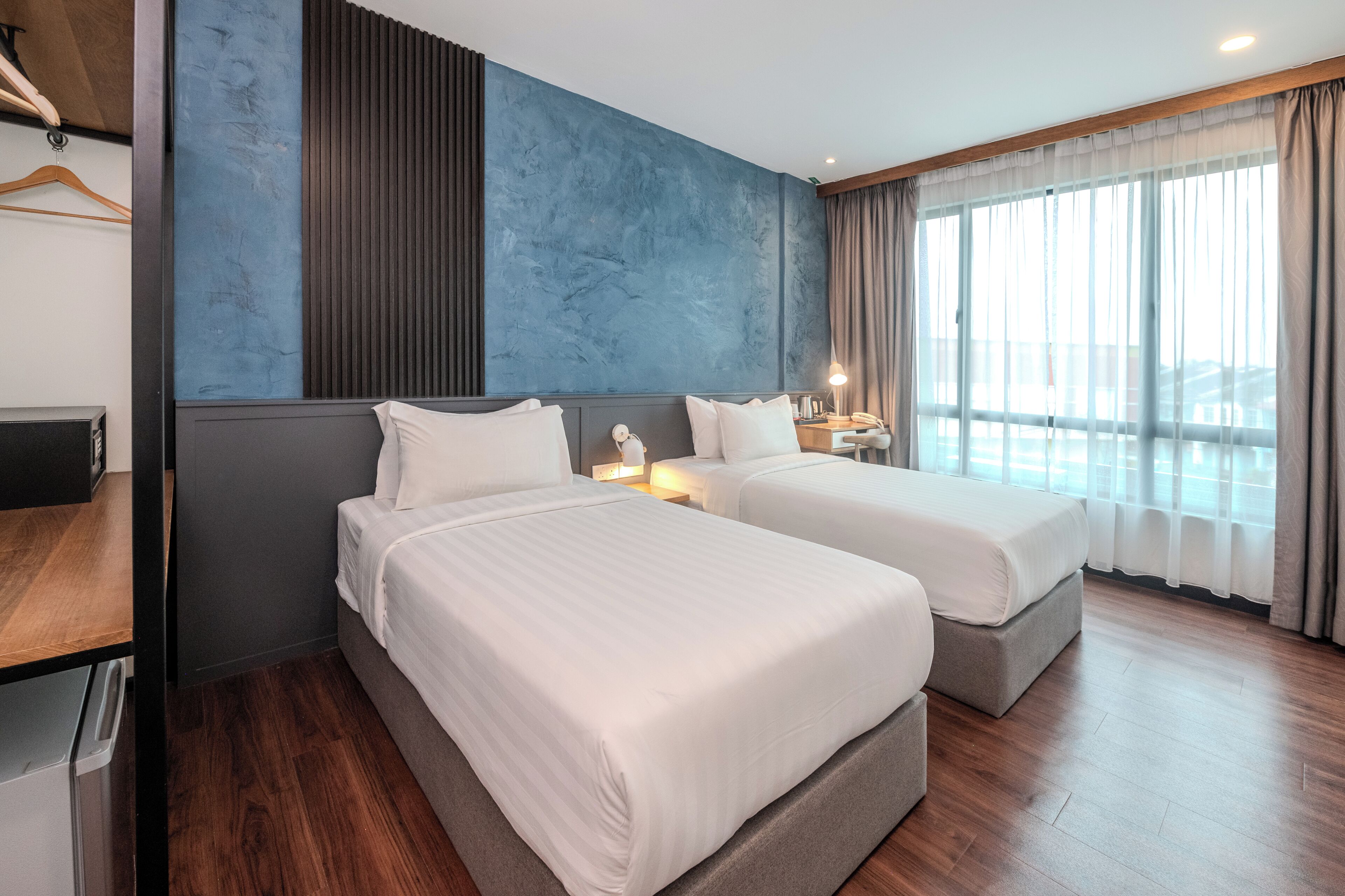 A hotel room with two beds, a wooden floor, a blue accent wall, and a large window with curtains.