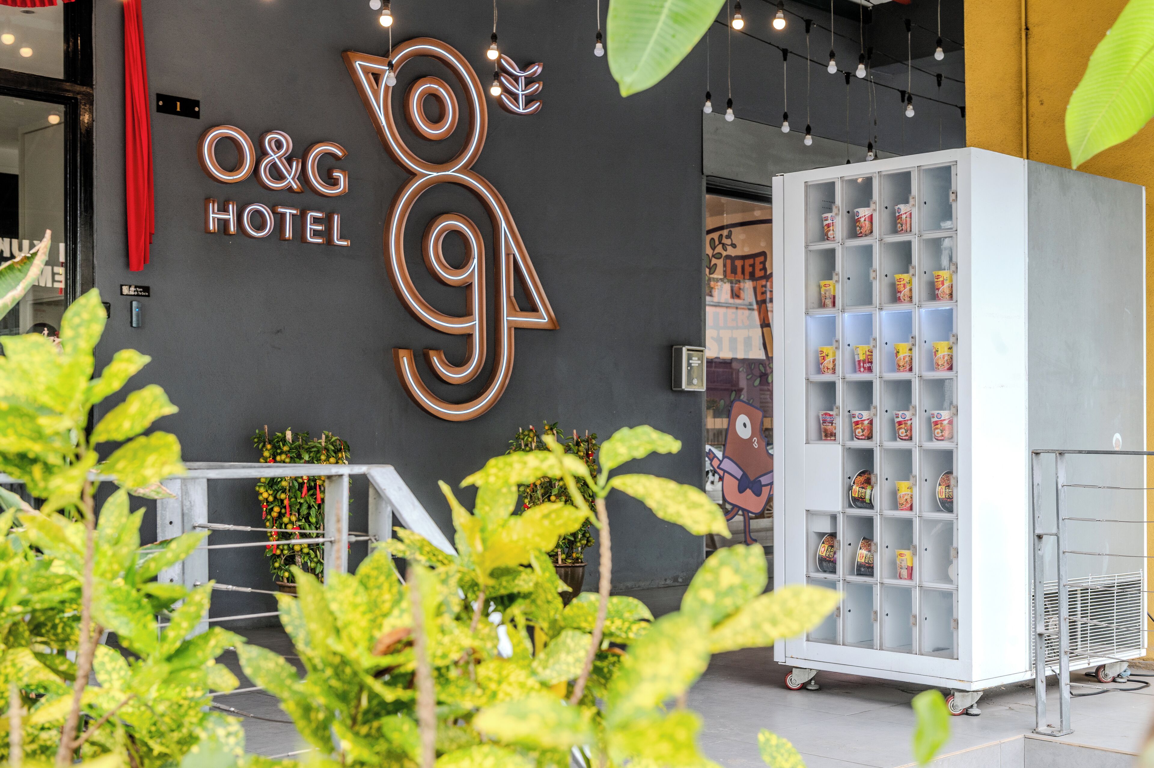 A hotel entrance with a neon sign, a vending machine, and potted plants.
