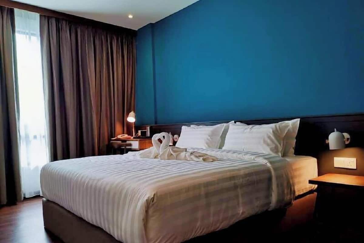 A hotel room with a large bed, a nightstand with a lamp, and a view of the outdoors through a window with curtains.