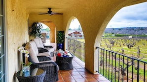 Eine überdachte Terrasse mit Rattanmöbeln, einem Deckenventilator und Blick auf einen Weinberg.