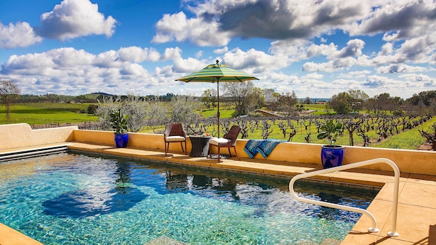Ein Schwimmbecken mit klarem Wasser, umgeben von einer gefliesten Terrasse, zwei Stühlen, einem Tisch und einem blauen Sonnenschirm.