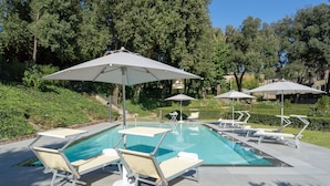 Piscina con sillones de descanso y sombrillas, rodeada de vegetación y árboles.