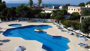 A swimming pool with multiple sections, surrounded by lounge chairs and umbrellas, with a view of the sea and buildings in the background. - Happy Days (Rhodes)