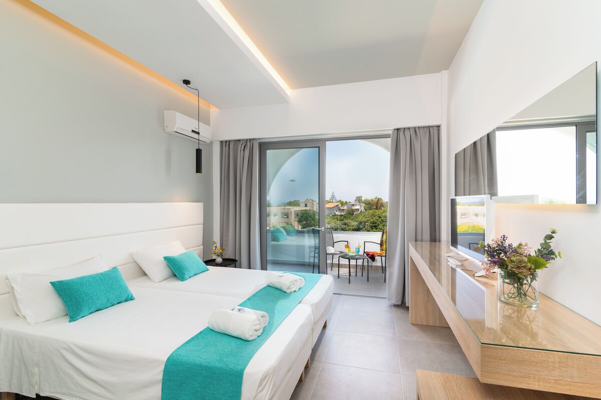 Habitación de hotel con cama, escritorio, televisor y balcón con vistas.