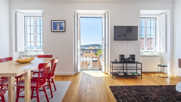 A spacious room with a wooden floor, a dining table with red chairs, a TV mounted on the wall, and a balcony with a view of buildings. - LV Premier Bica- BI (Lisbon)