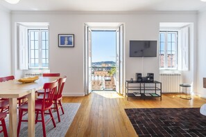A spacious room with a wooden floor, a dining table with red chairs, a TV mounted on the wall, and a balcony with a view of buildings. - LV Premier Bica- BI (Lisbon)