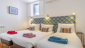 A hotel room with two beds, each with white linens and colorful striped pillows. There are towels in red, pink, and blue on the beds. The room has a window with a view of the sky and a wall-mounted air conditioning unit. - LV Premier Bica- BI (Lisbon)