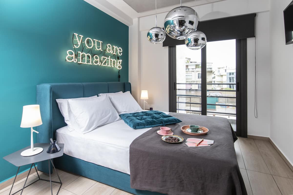 A modern bedroom with a teal accent wall, a bed with white and teal bedding, a bedside table with a lamp, and a neon sign.