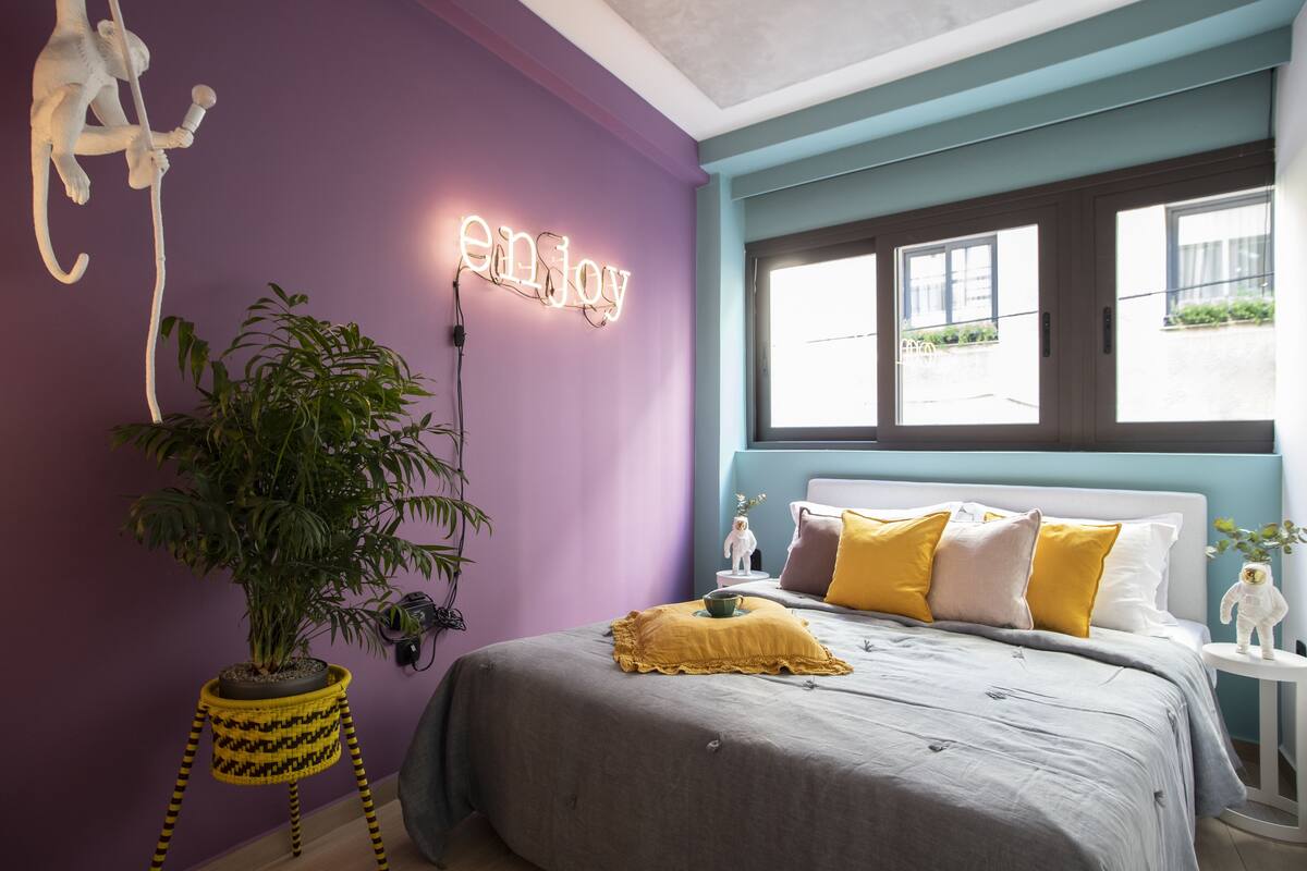 A bedroom with a bed, a potted plant, a wall-mounted light, and a window.