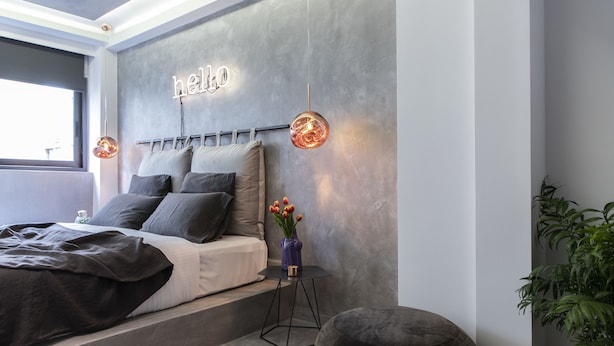 A modern bedroom with a bed, a bedside table with a vase of flowers, a wall-mounted light fixture, and a 'hello' sign. - 14 Reasons Why (Athens)
